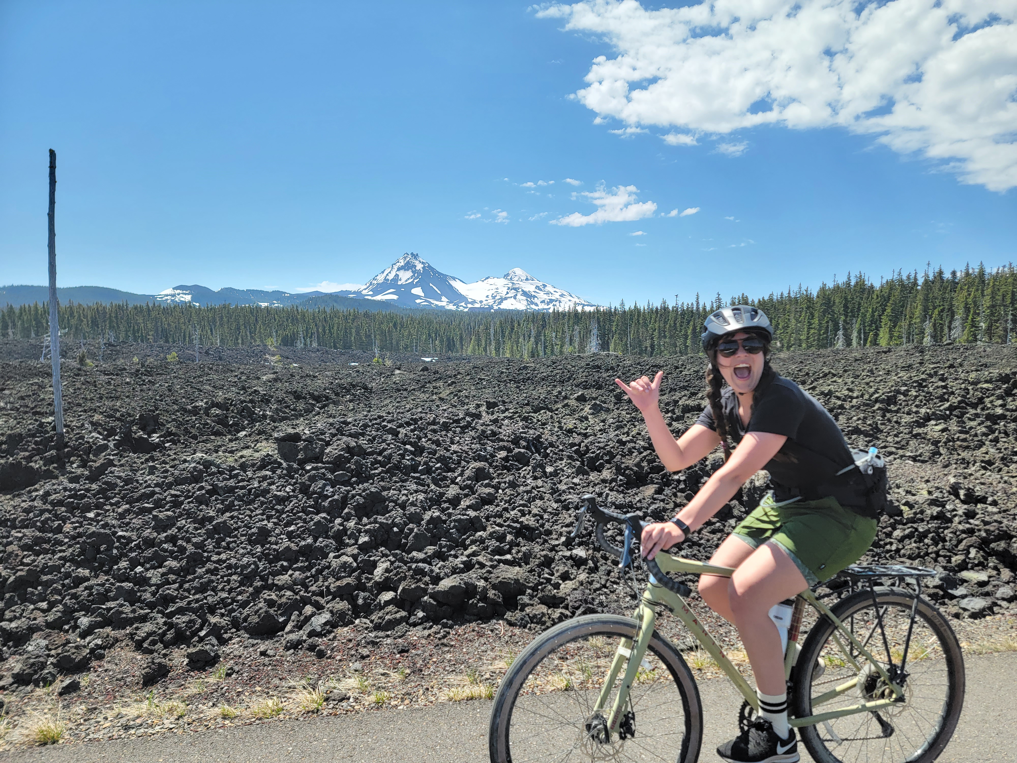 A picture of a woman biking up the McKenzie Pass, looking at the camera and giving a hang loose, with lava rock and mountains in the background